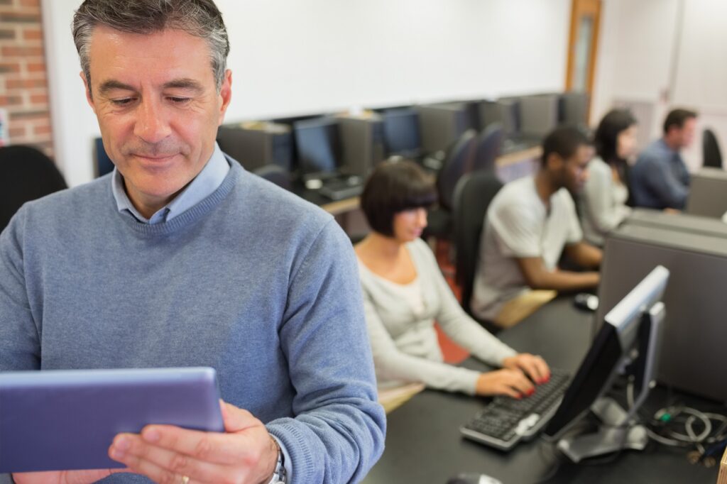Teacher standing holding a tablet PC in computer class in college